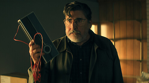 Older man with glasses and a beard stands indoors in a warmly lit room, holding up a device with a red wire, wearing a dark plaid shirt and jacket, looking intensely at something off camera.