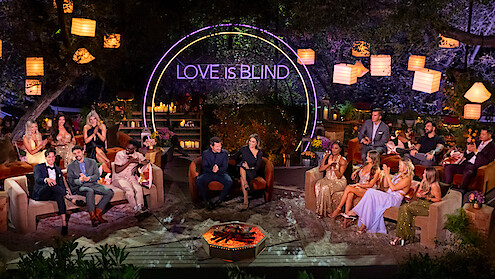 A group of well-dressed people sit in a semi-circle outdoors at night, under lanterns and trees, with a neon "Love Is Blind" sign behind them, creating a festive, lively, and glamorous TV show reunion atmosphere.