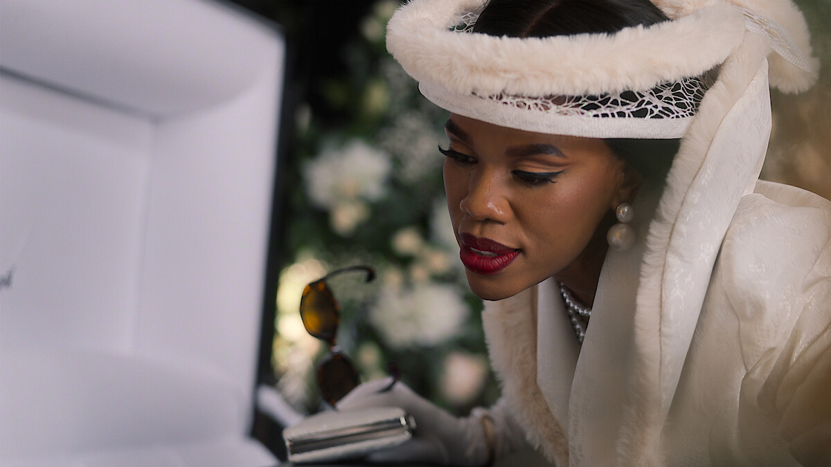 Woman in elegant white attire with pearls and fur hat looking into an open casket at a funeral, holding sunglasses and a clutch, with floral arrangements in the background.