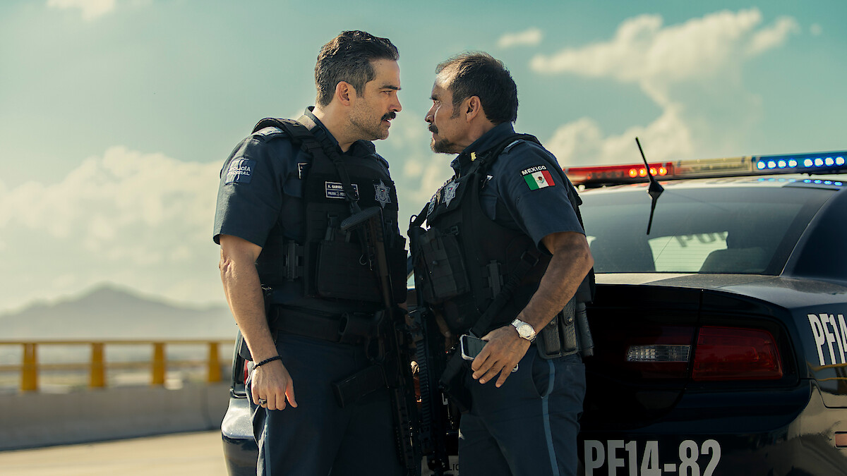 Dois policiais uniformizados frente a frente em um confronto tenso ao lado de uma viatura com as luzes acesas durante o dia, com montanhas e o céu azul ao fundo.