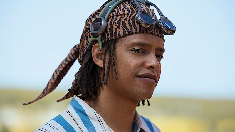 Jacob Romero as Usopp wearing a striped shirt, tiger-patterned headscarf, and goggles stands outdoors in a sunny natural landscape with a blurred background.