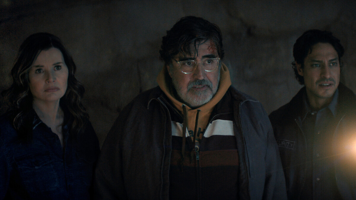 Three people stand together in a dimly lit, cave-like environment, looking ahead with serious expressions. One person holds a light source, adding a dramatic glow to the mysterious scene.