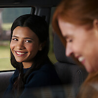 Two women sitting together in a car, one smiling warmly at the other; the environment appears relaxed and friendly, with natural daylight coming through the window.