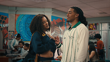 Two young adults talking closely in a colorful school cafeteria, with other students sitting and interacting in the background, murals and bright artwork on the walls.