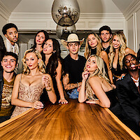 Group of young adults posing around a long wooden table in an elegant, modern indoor setting with large pendant lights above and white walls in the background.