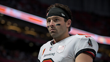 Football player in a white Tampa Bay Buccaneers uniform with black eye paint and headband, standing on the field in a stadium with a blurred background and stadium lights.