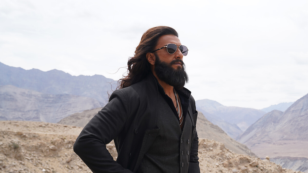 Man with long hair and beard wearing sunglasses and dark clothing stands confidently in a rugged mountainous landscape under a cloudy sky, suggesting a cool and stylish vibe in a remote outdoor environment.