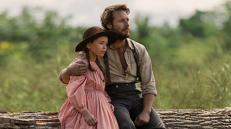 A man and a young girl in old-fashioned clothing sit on a large log outdoors. The man has his arm around the girl, and they both look thoughtfully into the distance, surrounded by greenery and soft natural light.