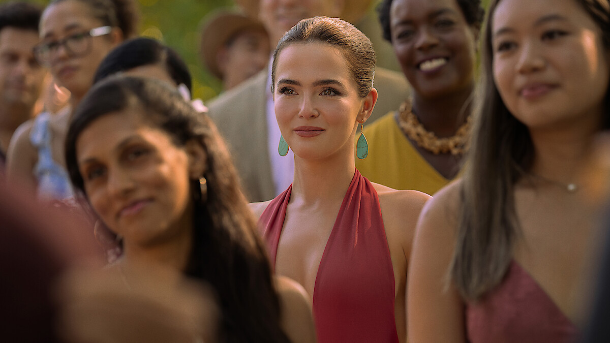 A young woman in a red dress with turquoise earrings stands among a diverse group of smiling people outdoors in warm daylight, suggesting a happy social event or celebration.